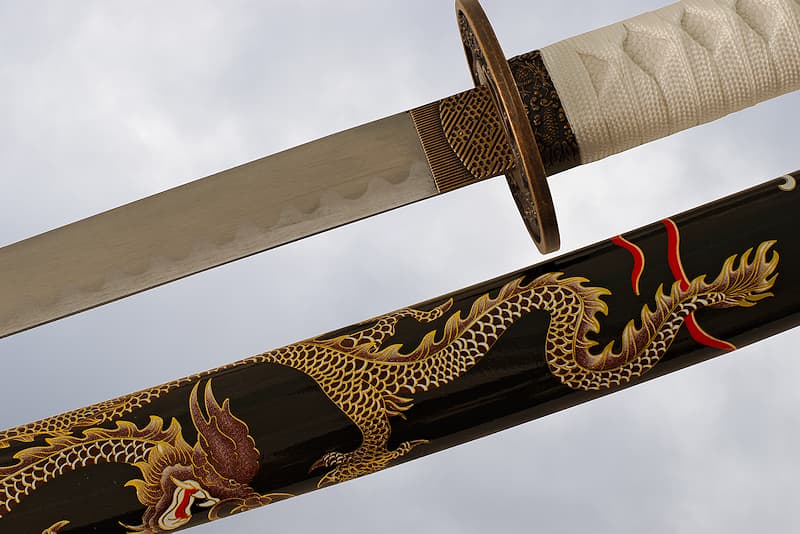 Collector's Katana with black saya (鞘) decorated with a screen-printed dragon, dragon tsuba (鍔), white tsuka-ito (柄糸) and sageo (下緒) | Legend has it that I was forged in the belly of the Celestial Dragon. The master blacksmith, wanting to save his son who had been eaten alive by the animal, took the same route. After finding his son, he set out to find the materials swallowed by the monster to forge a blade.