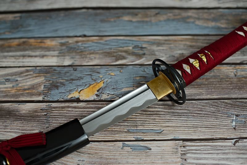 Sharp wakizashi, damascened forge, with black scabbard (saya 鞘), tsuka-ito (柄糸) and dark red sageo (下緒), delivered in a wooden box containing a maintenance kit and a protective cloth cover | It was in a bath of blood that I was born. Renowned but discreet, the blacksmith who gave birth to me was a victim of his own success. Brigands robbed him just as he and my sister were finishing forging my katana blade. Having no mount at the time, they paid no attention to me on the polishing stand. But when the samurai master to whom I was destined discovered the forfeit, he found a new craftsman to create my blood-red mount.