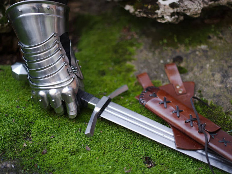 A knight's hand protected by an articulated gauntlet takes possession of a forged medieval sword resting on a moss-covered rock.