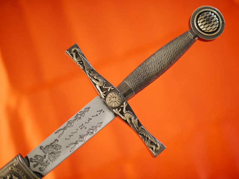 Excalibur protected by its braided scabbard, placed on a medieval table decorated with vegetation and a few dewdrops. Excalibur, King Arthur's sword, with red braided scabbard #Terressens