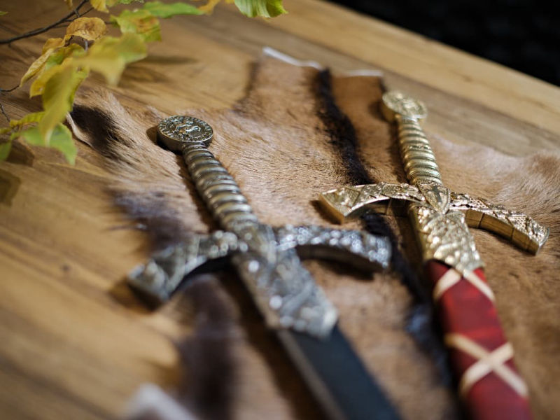 The two decorative Templar swords with red braided or black scabbards, placed on a medieval wooden table, with an autumn branch. The two decorative Templar swords with red braided or black scabbards, placed on a medieval wooden table, with an autumn branch.