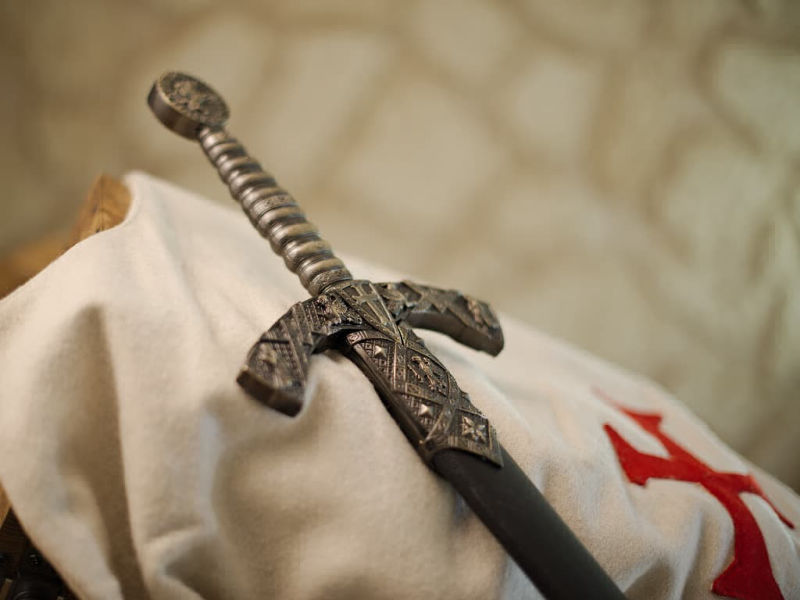 Decorative Templar sword with black scabbard, set against a white Templar tabard bearing the red Templar cross. Decorative Templar sword with black scabbard, set against a white Templar tabard bearing the red Templar cross.