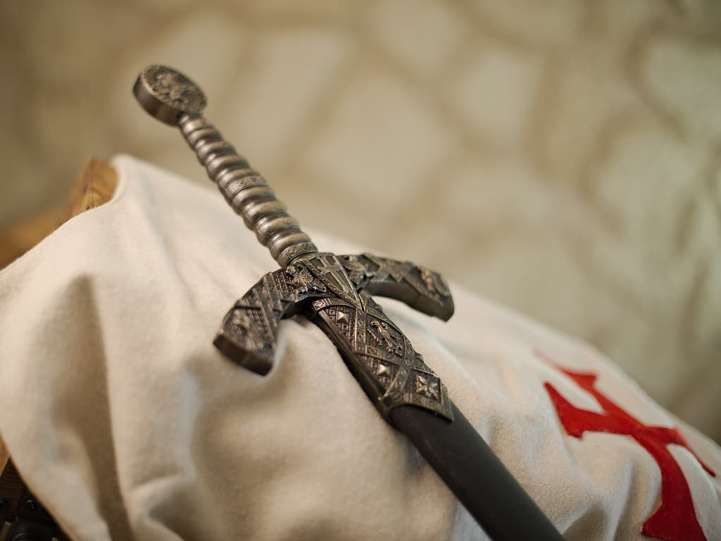 Decorative Templar sword with black scabbard, set against a white Templar tabard bearing the red Templar cross. Decorative Templar sword with black scabbard, set against a white Templar tabard bearing the red Templar cross.