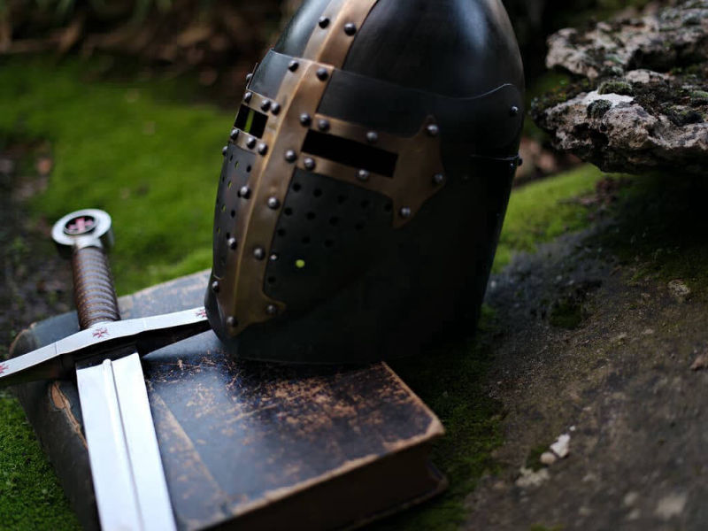 Templar black helmet with brass cross, placed on a sword and an ancient book.