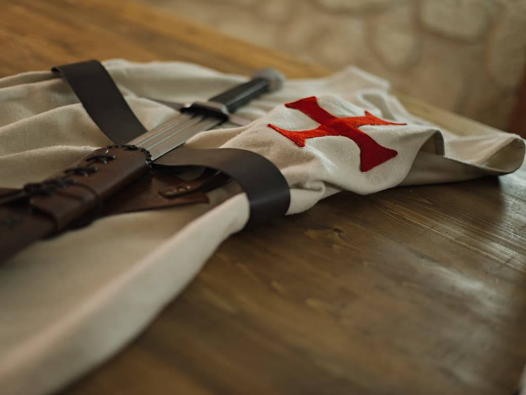 Tabard templier avec épée médiévale de combat posés sur une table traditionnelle en bois Tabard templier avec épée médiévale de combat posés sur une table traditionnelle en bois